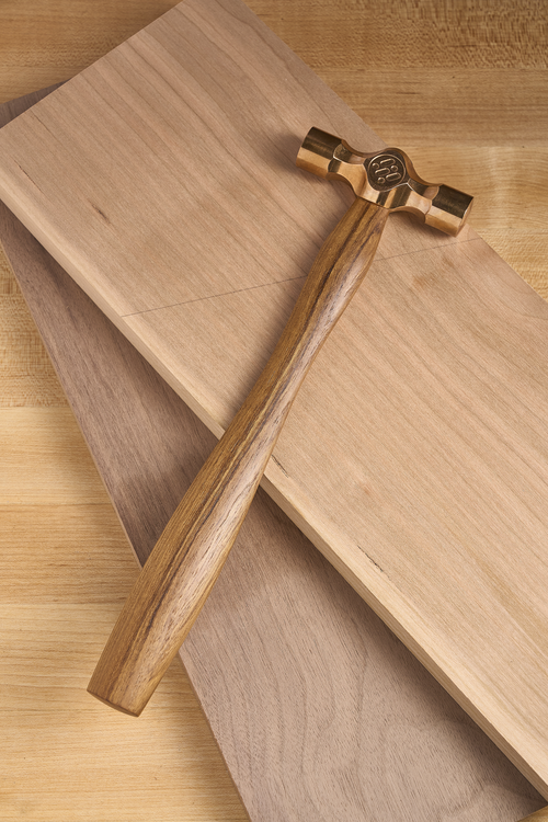 Cross peen hammer with brass head and wooden handle resting on wood boards